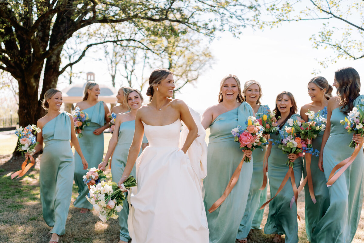 Bride walking and laughing with her bridesmaids in light blue dresses while holding colorful bouquets on the lawn at Burritt on the Mountain in Huntsville, Alabama, photographed by Perry Hancock.