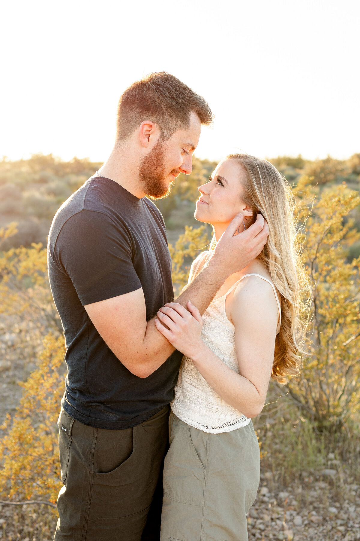 A couple gazes lovingly into each other's eyes during golden hour in the desert, the soft sunlight highlighting their expressions and the natural landscape behind them.