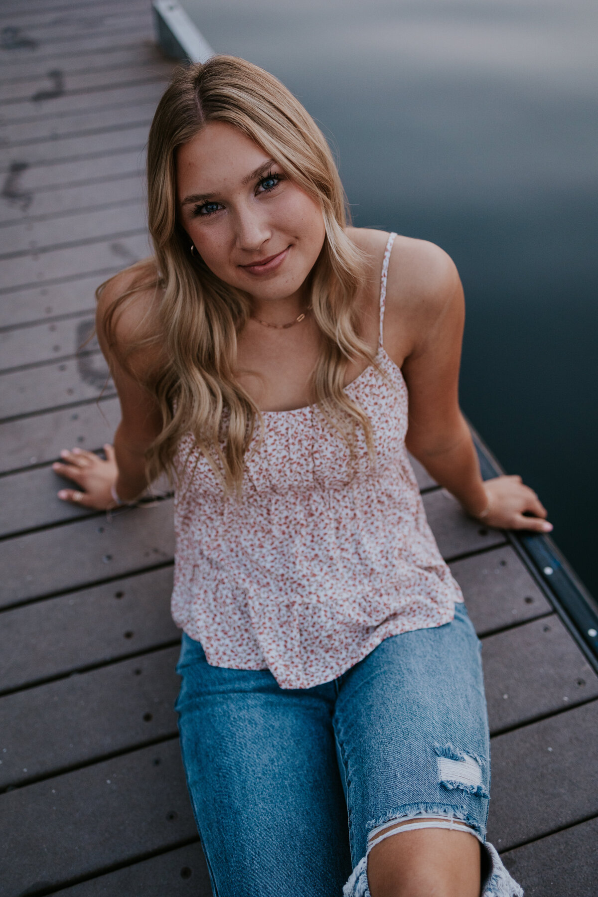 Young woman sits on dock in summer clothes while looking up at camera.