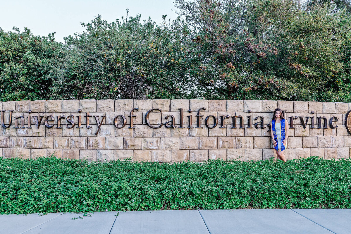 A person stands next to a large stone wall with University of California, Irvine inscribed on it. Lush greenery surrounds the base of the wall, and trees are in the background. The person is wearing a blue outfit.