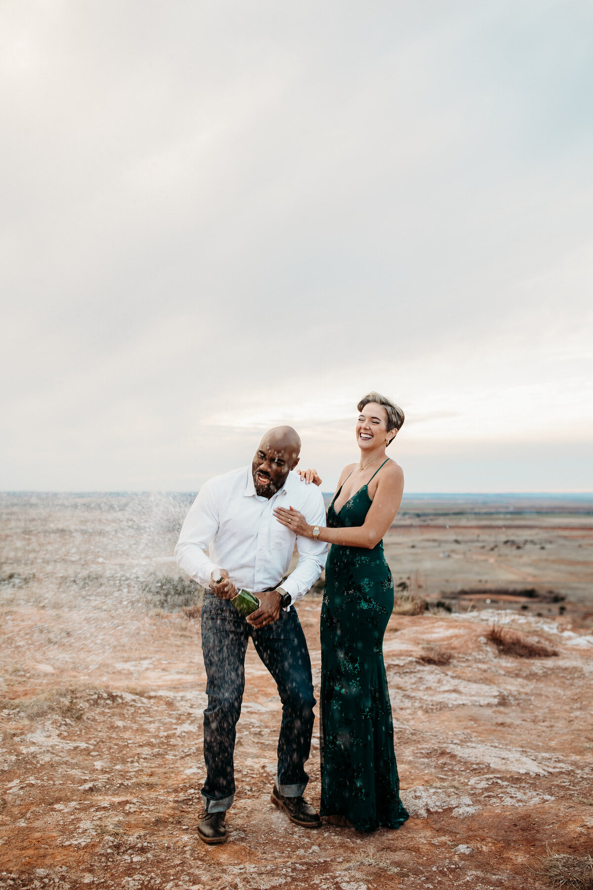 A couple laughing while standing on a rocky field spraying champagne.