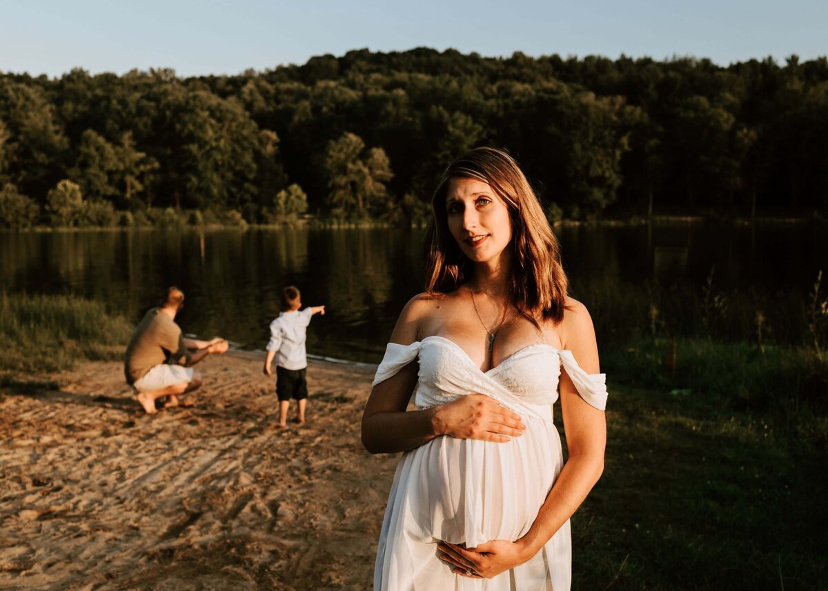 A pregnant woman standing in front of a lake with her family captured by a Pittsburgh maternity photographer.