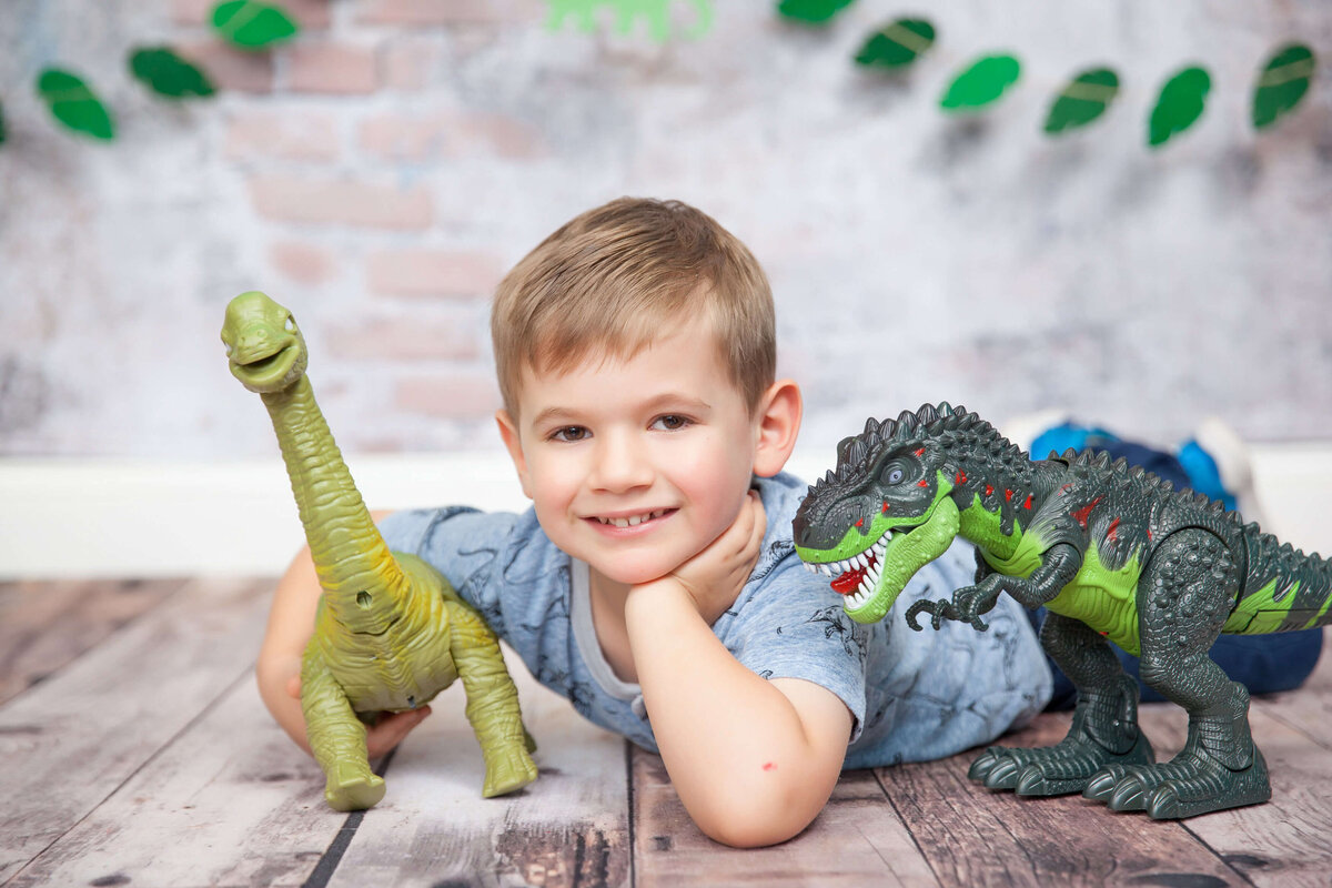 A young boy with short blonde hair is lying on the floor with his head resting on his hands, smiling at the camera. Flanked by two toy dinosaurs—one a Brachiosaurus and the other a Tyrannosaurus rex—he's captured in this charming moment by Jacksonville family photography against a faux brick wall backdrop.