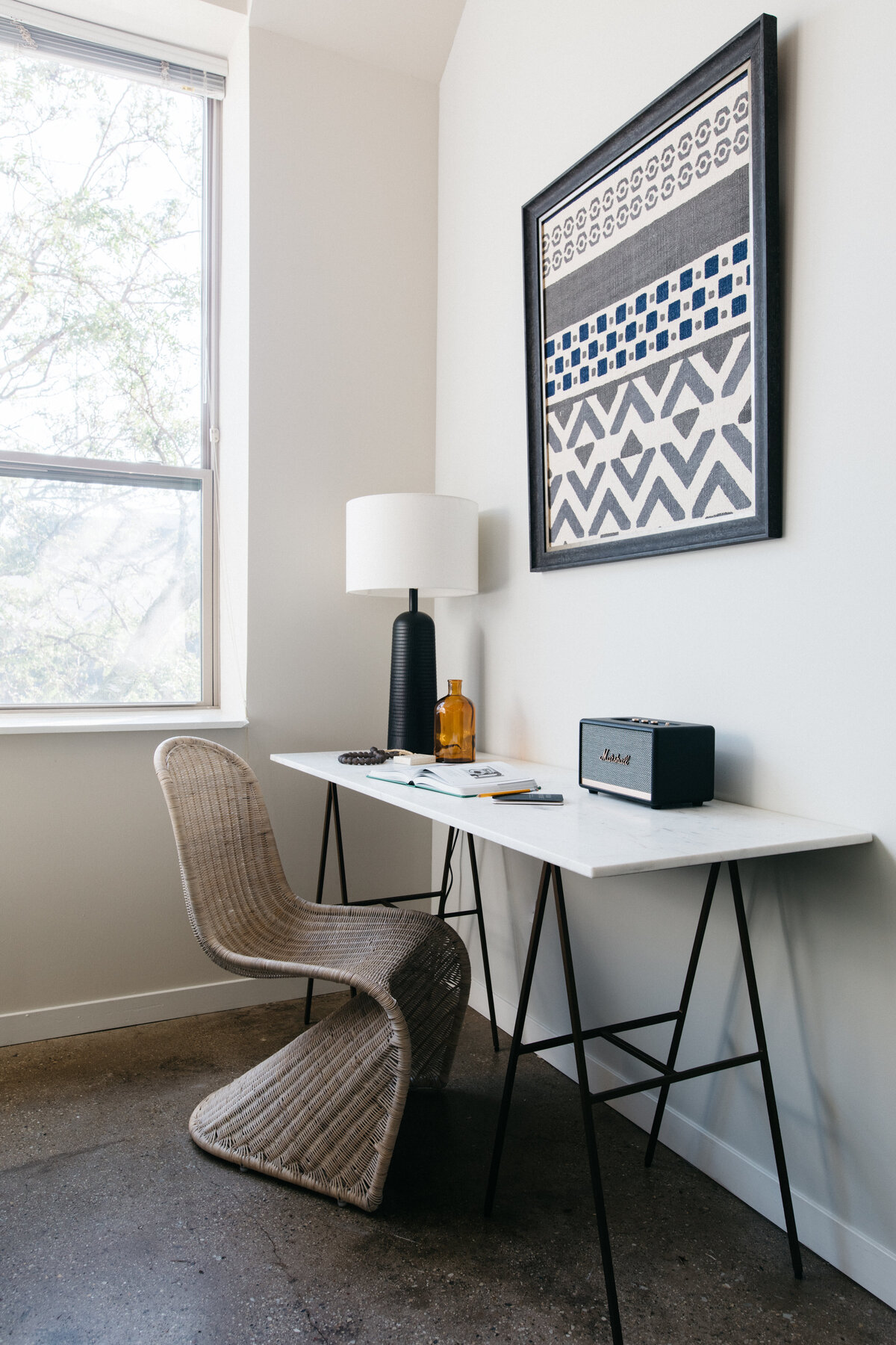 Minimalistic home office with marble trestle table as desk, wicker saarinen style chair, black ceramic table lamp and black and white geometric art on wall