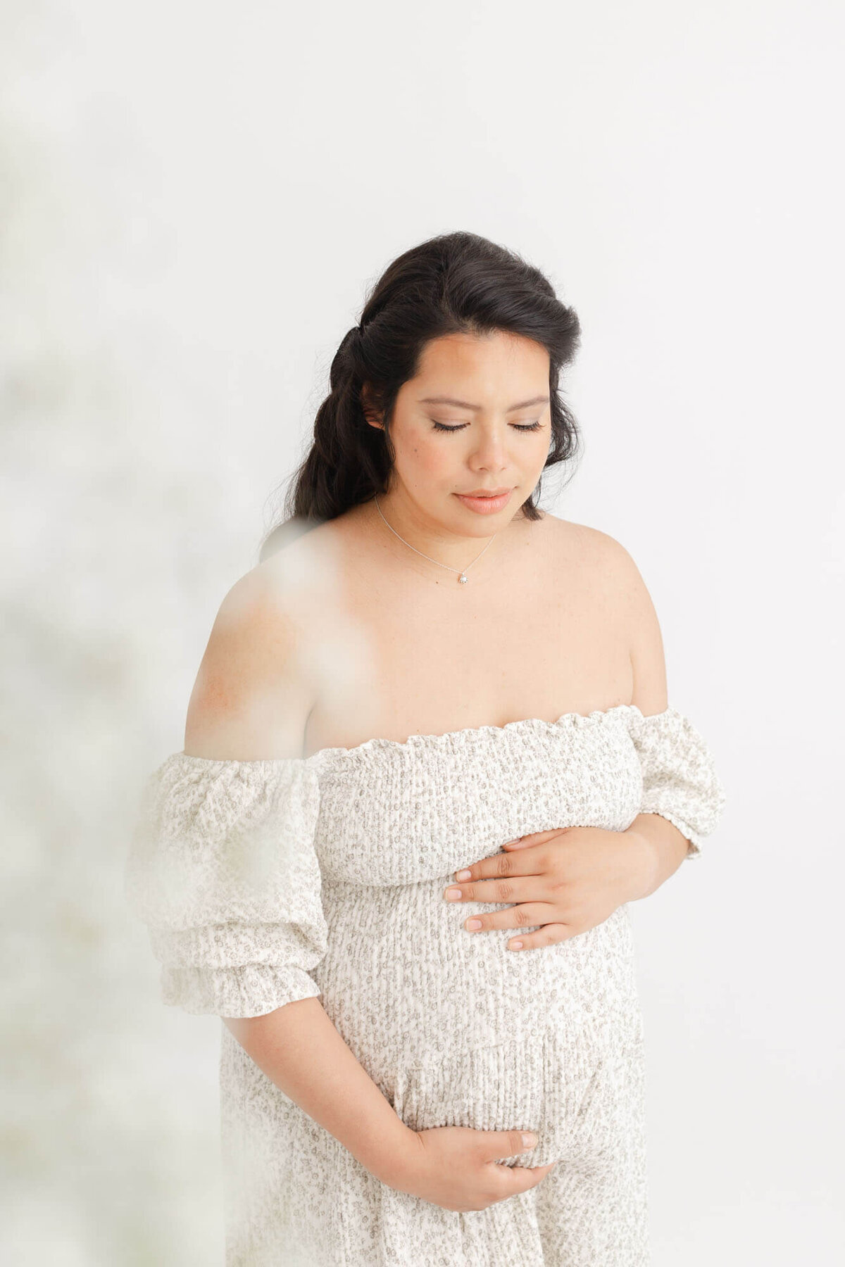 Pregnant Woman holding belly and looking down with a soft grin on her face. There are some white babys breath flowers in the foreground.