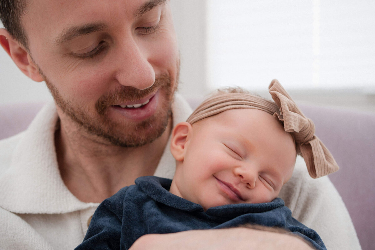 Father smiling while holding a newborn baby wearing a headband, who appears to be smiling in their sleep.