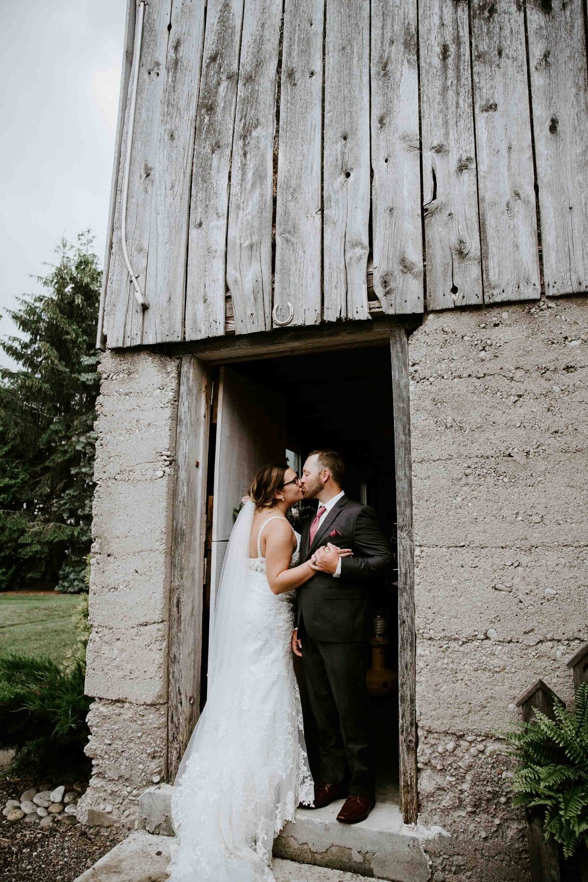 Bride and groom are standing in the doorway of a rustic barn in Exeter, Ontario and kissing for a posed wedding day photo.