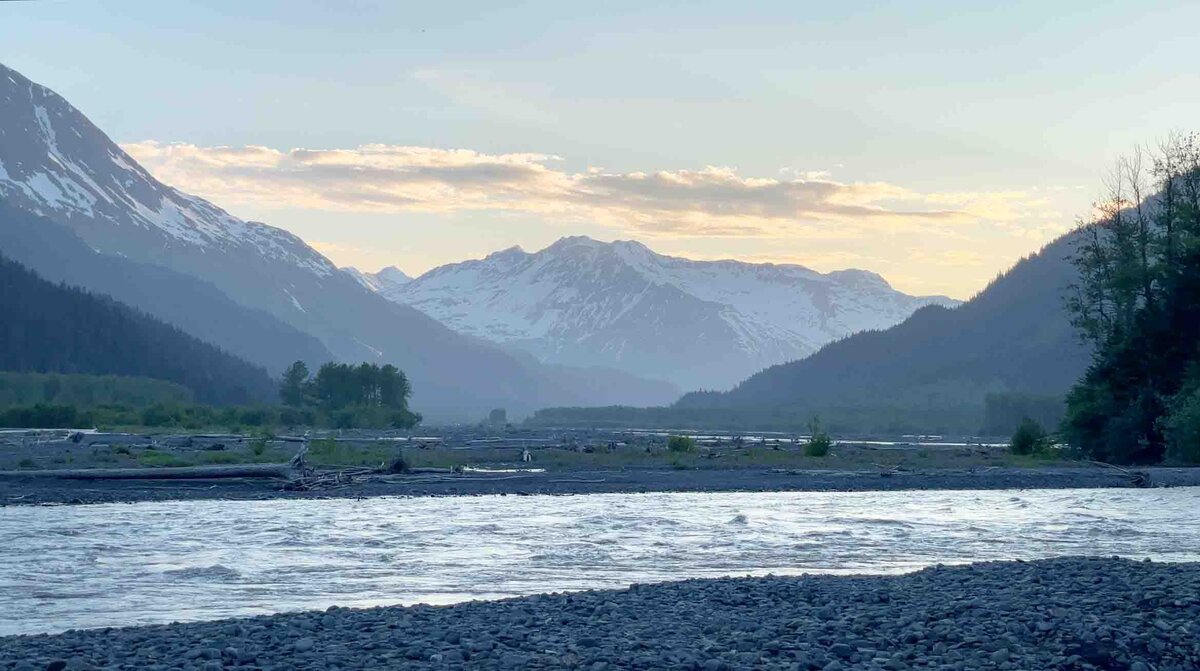 A view of craggy snow covered mountains, a river flowing and a rocky shoreline bathed in sunset glow during an elopement wedding with guests in Denali National Park as part of an adventure luxe elopement package in Alaska. 