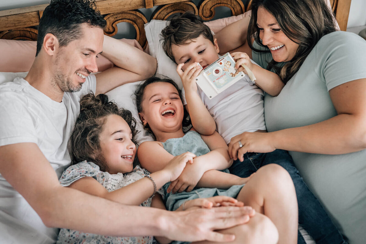 A family of five lies on their bed, tickling and laughing, during an in home maternity photoshoot.