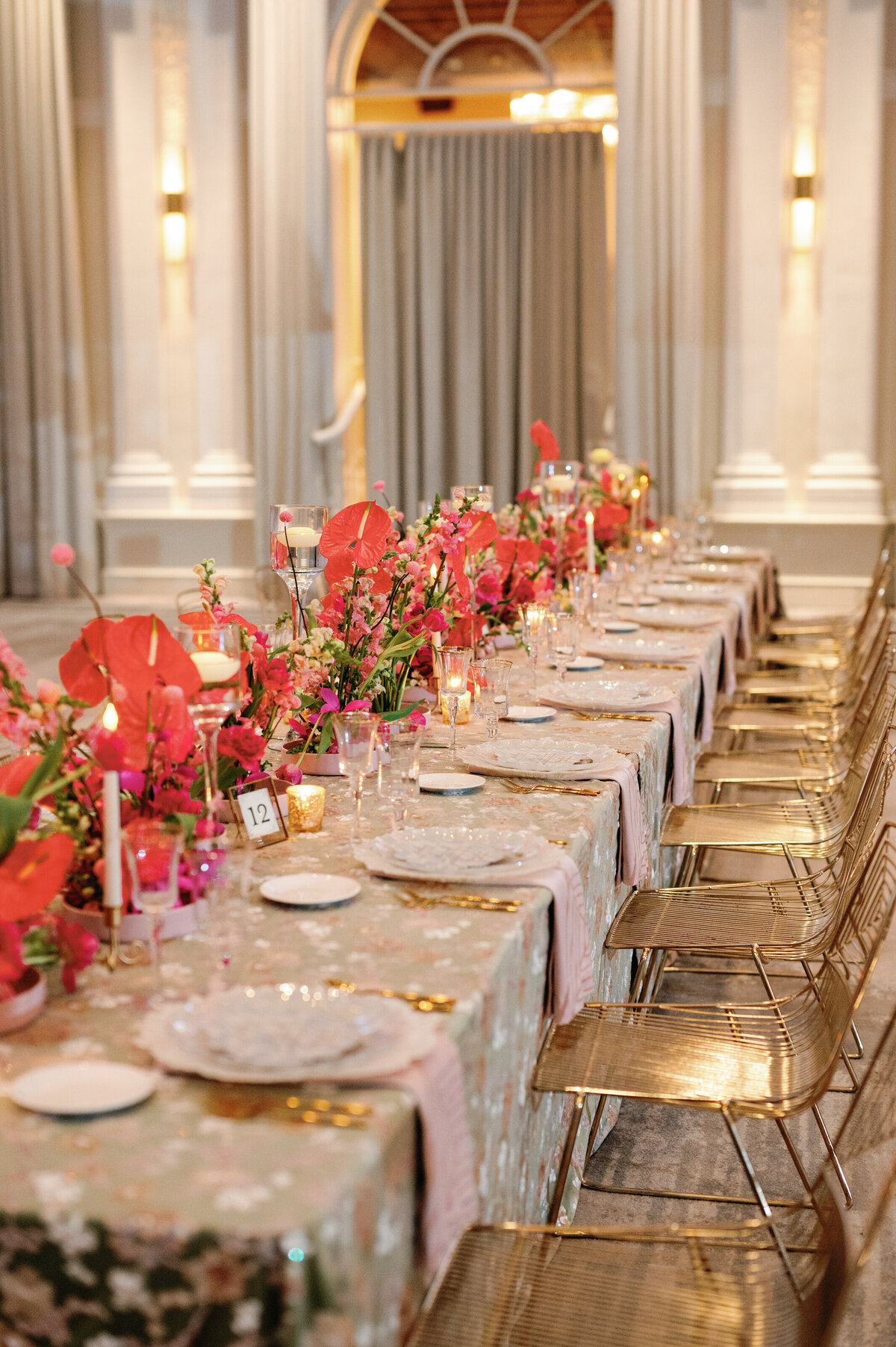 An exquisite wedding tablescape featuring gold chairs, elegant gold utensils, and frosted white dishes, beautifully set atop a floral-patterned tablecloth. The pink-themed floral centerpieces add a romantic and sophisticated touch, creating a timeless and luxurious reception setting.