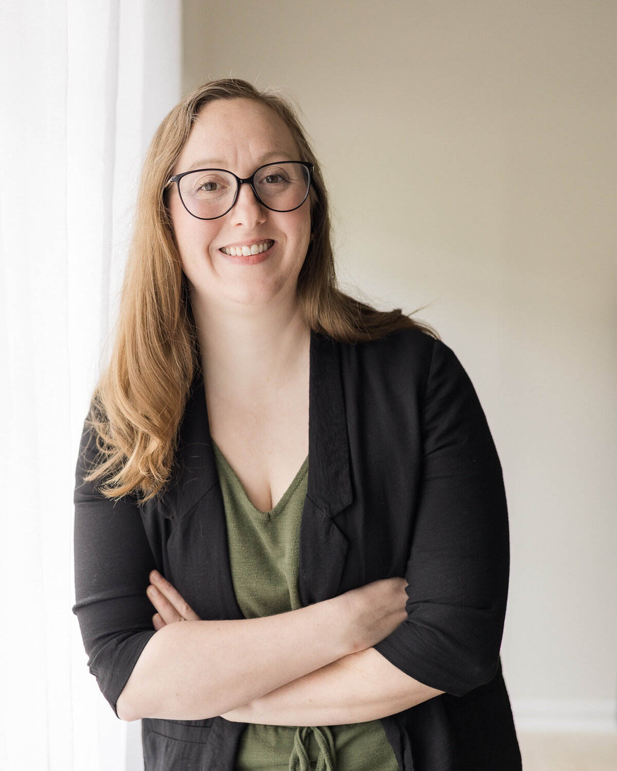 Strawberry blonde female, standing with her hands crossed leaning against a window, wearing a dark green shirt with a black jacket over the green outfit, wearing glasses, smiling at the camera