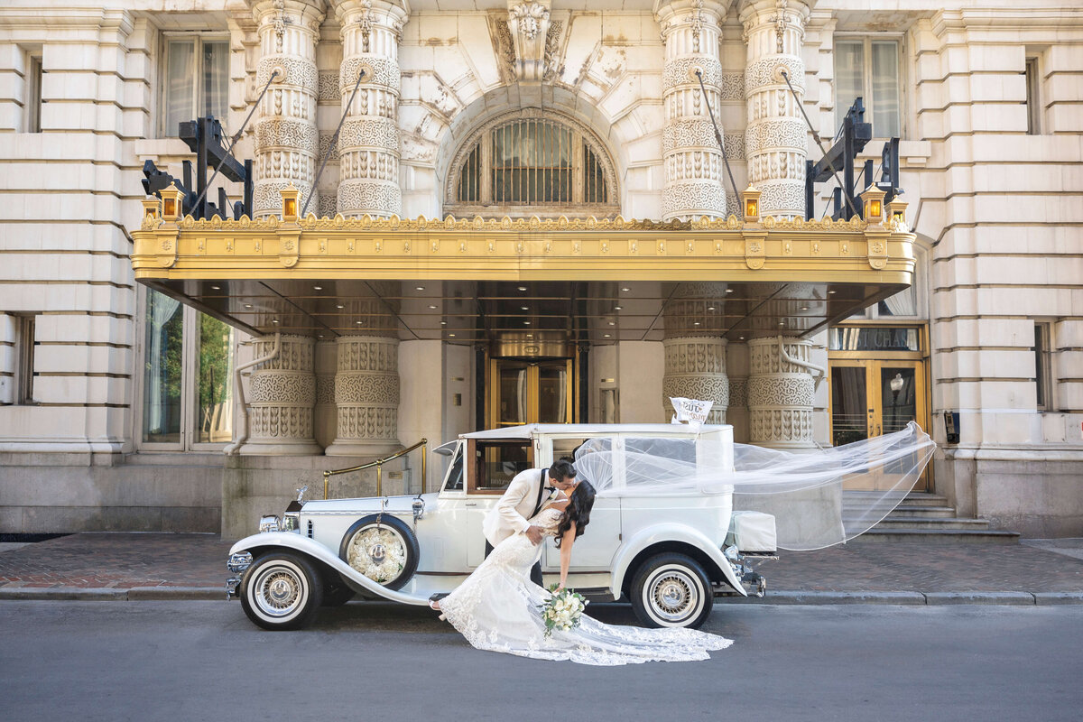 A bride and groom pose in front of a vintage white car with a flowing veil draped dramatically. The backdrop is an ornate building with intricate columns and a grand entrance, creating a classic and elegant wedding scene.