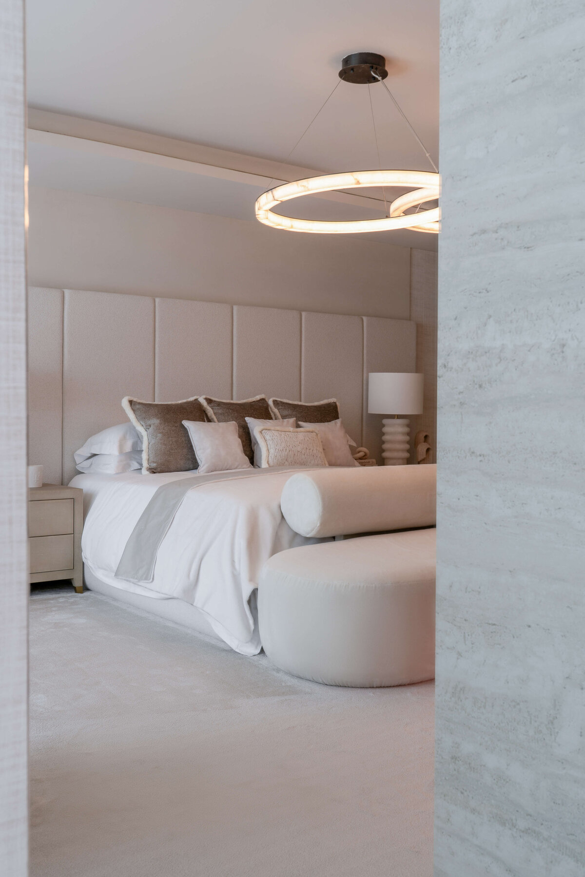 From outside the room, a glimpse into a spacious primary bedroom with an airy, neutral aesthetic . Above the large, white bed is a contemporary circular light fixture.