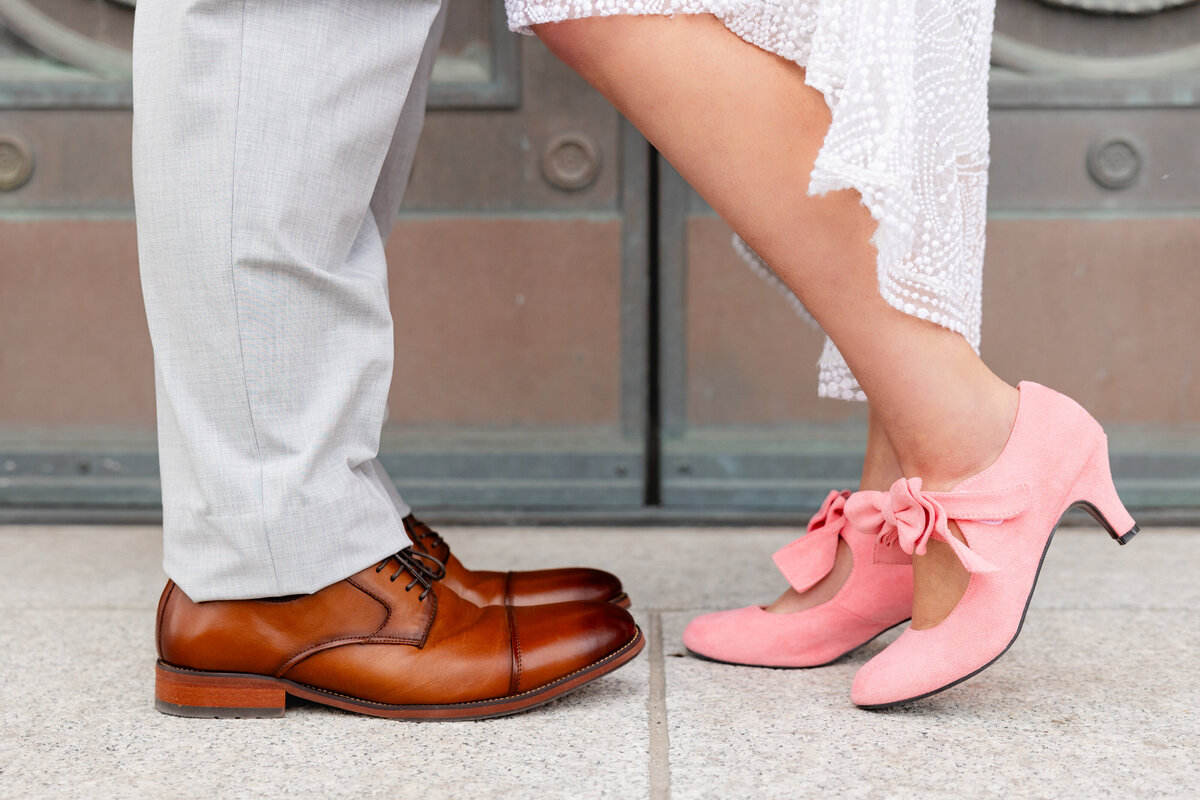 A close-up of the bride’s unique pink bow heels alongside the groom’s classic brown leather shoes, symbolizing their perfect contrast and connection.