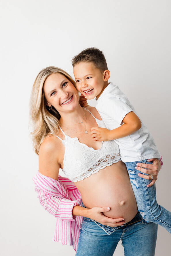 a pregnant mom holds her older son while touching her bare belly.