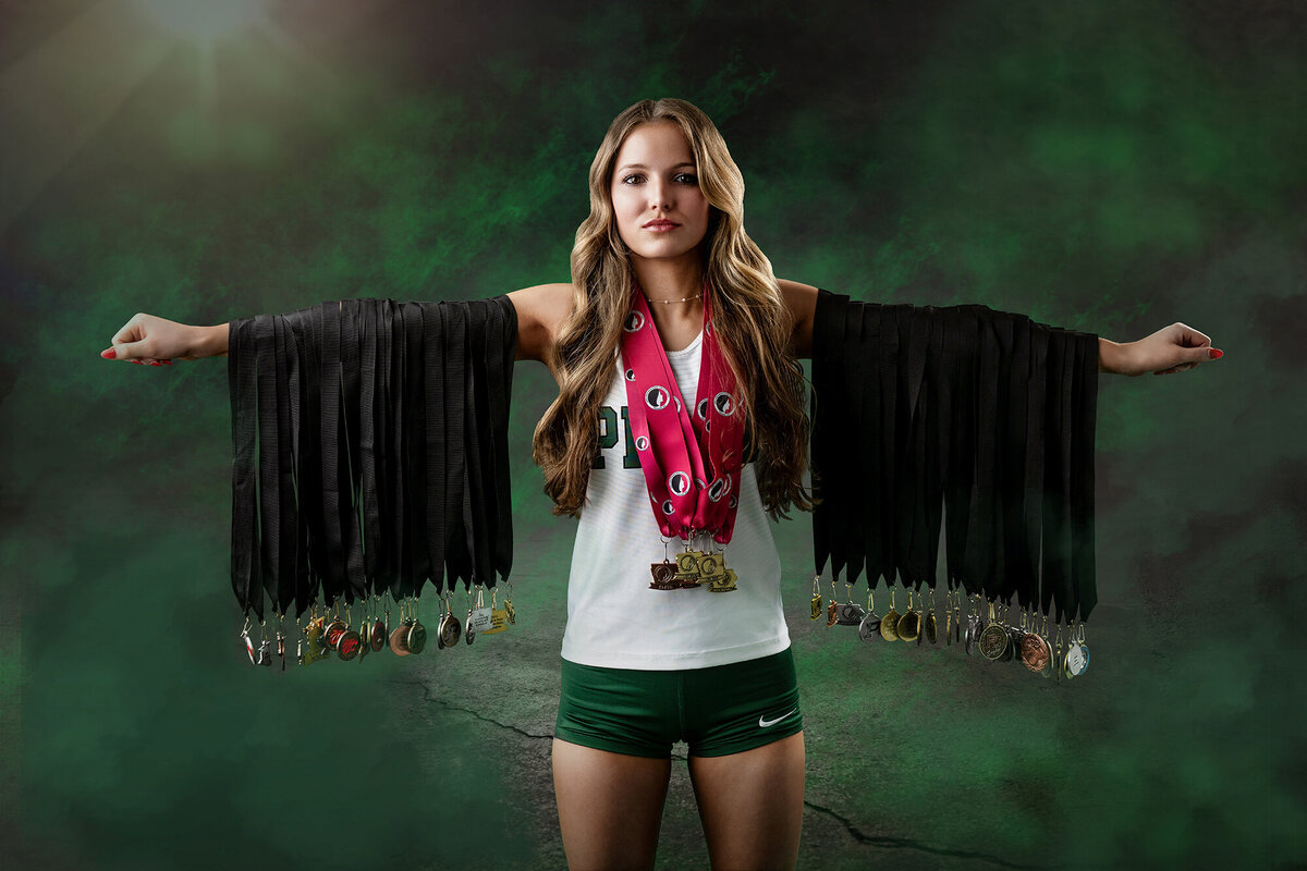 A high school senior stands in a studio showing off her many medals hanging from her extended arms and neck in her pella high school jersey in the studio of a Pella Senior Photographer