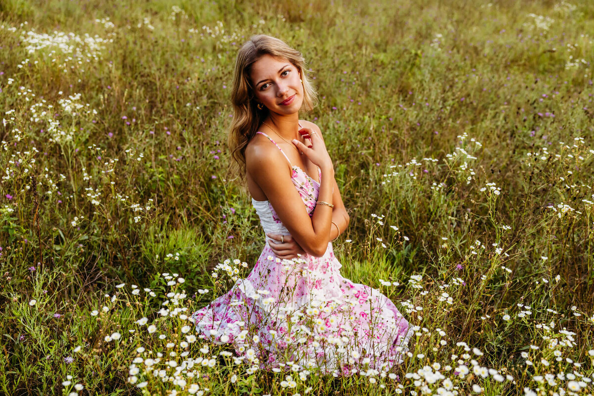 super pretty young lady in a pink and white floral dress kneeling in a flower field and playing with her necklace as she glances over her shoulder taken by Ashley Kalbus