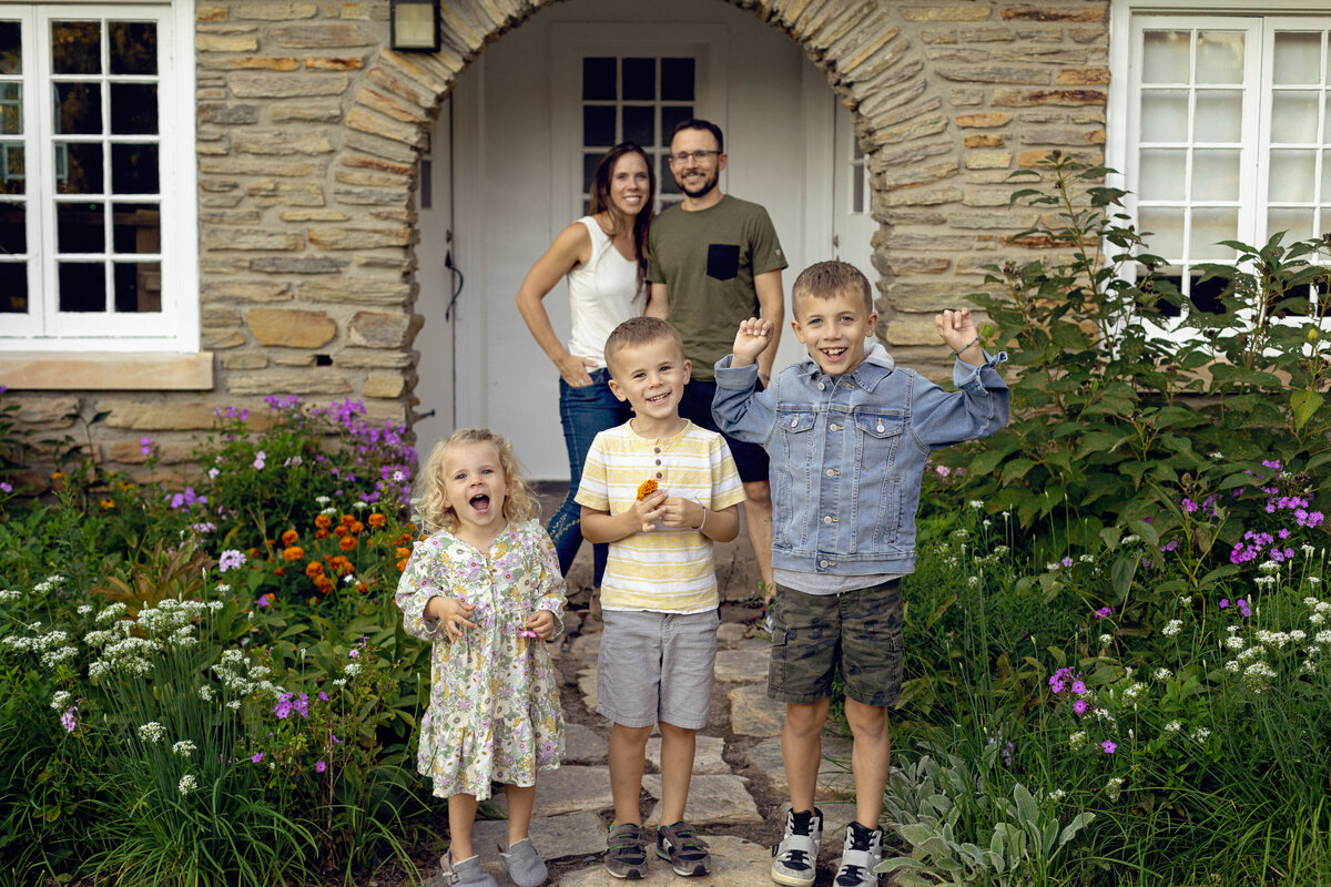Three siblings smile and stand at the entrance to their house with mom and ad standing on the porch as posed by a New Jersey Family Photographer