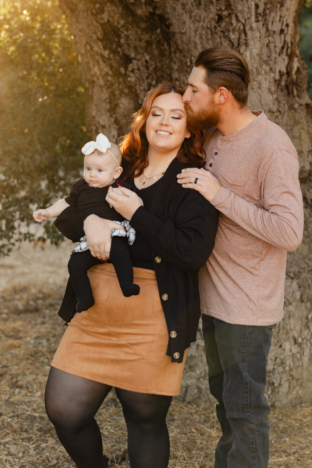 Mother sitting on chair holding baby while father stands close beside her during golden hour. Photographed by Yucaipa family photographer Kaitlyn Dawn Photography.