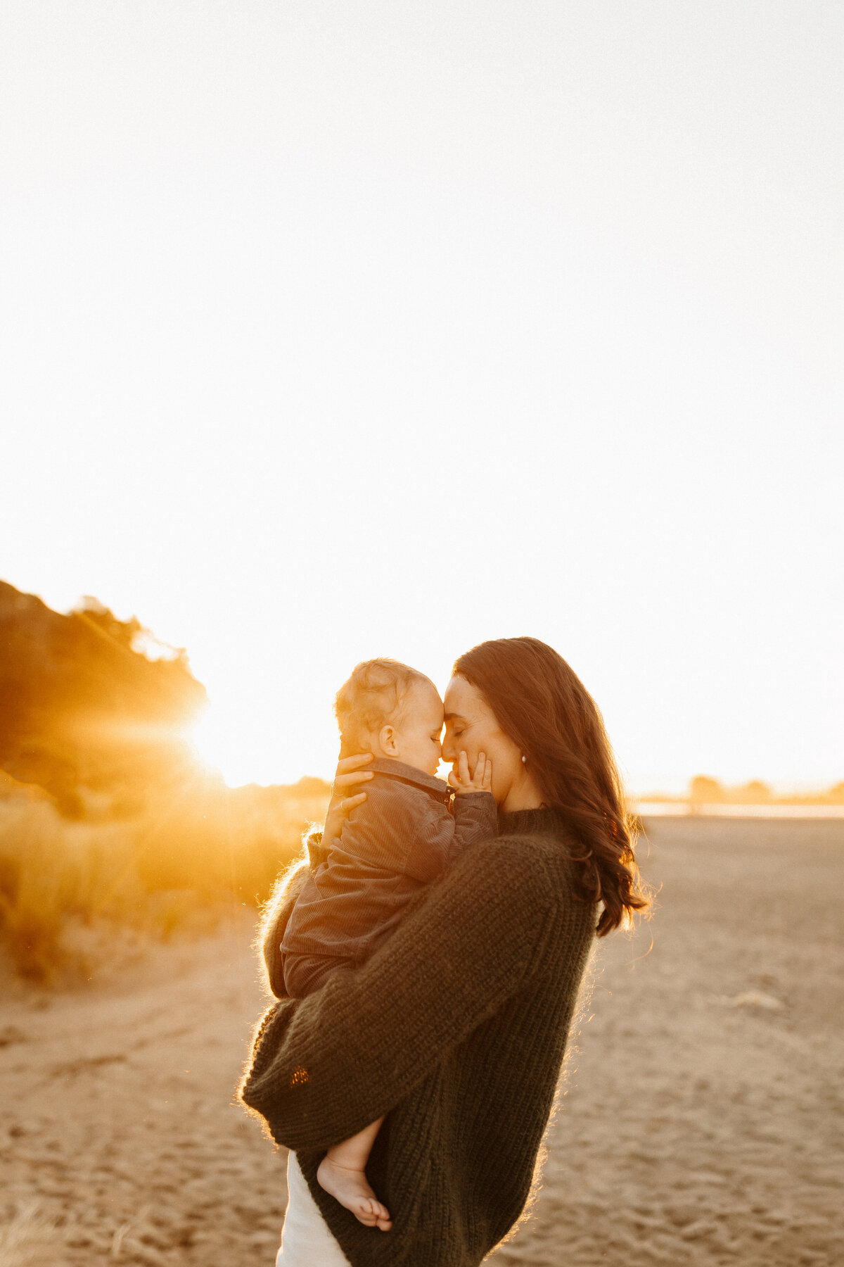 A mother holding her child in the golden glow of sunset while being photographed by lucy jane at a popular christchurch beach
