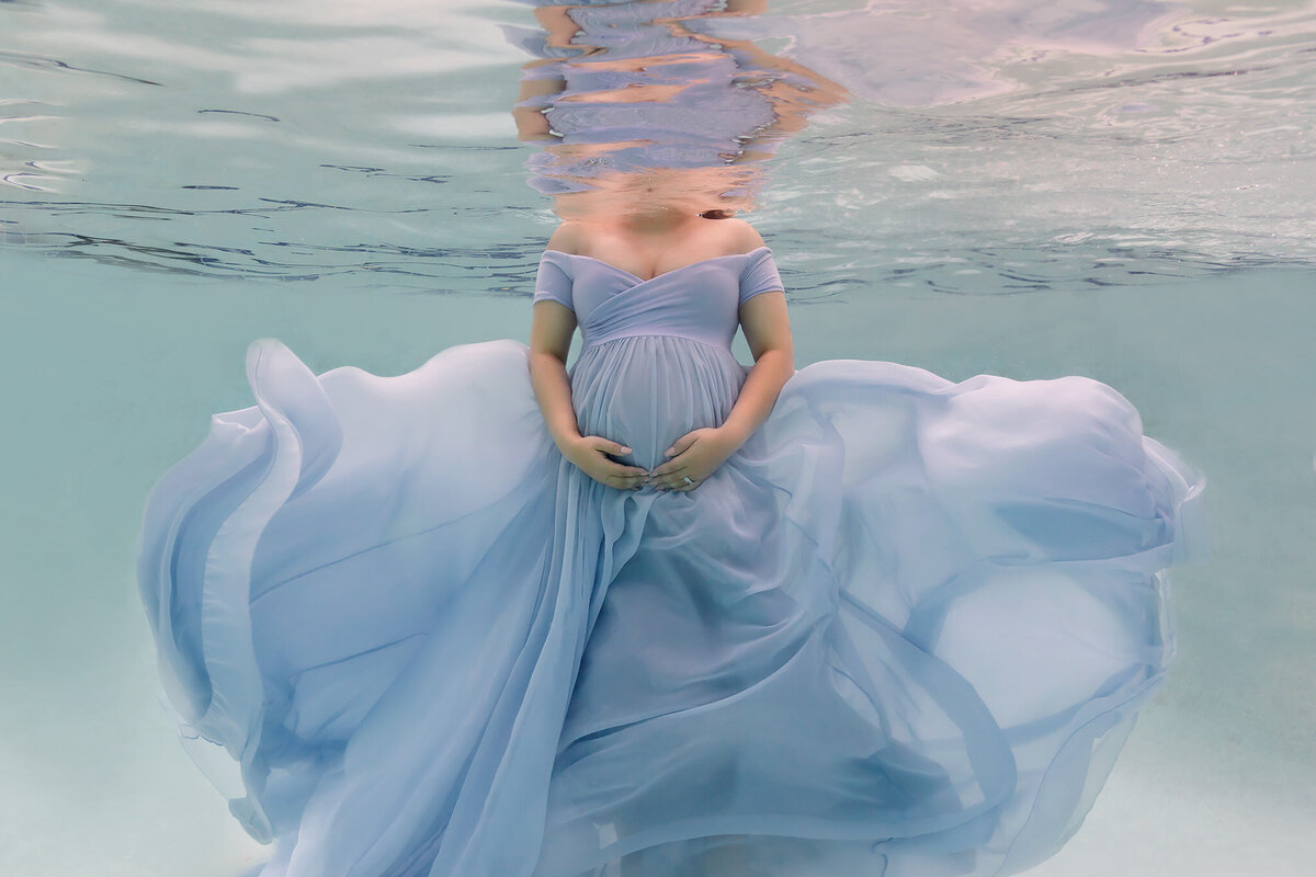 Underwater maternity photo capturing a woman cradling her belly while wearing a flowing blue gown. The soft, airy fabric swirls around her, enhancing the dreamy atmosphere created by this Dallas underwater maternity photographer.