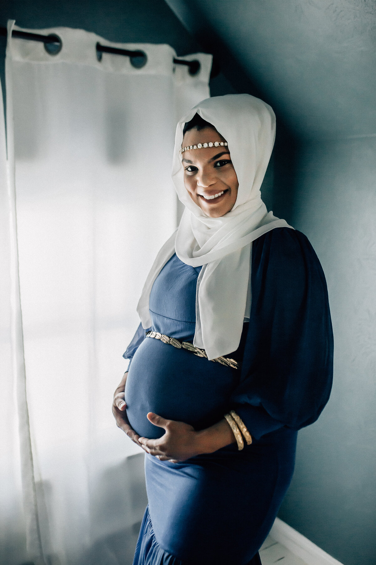 Maternity photoshoot with woman wearing blue dress, white scarf, and holding her belly.