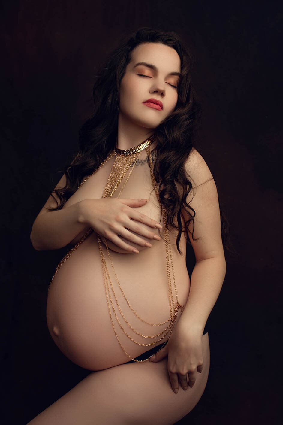 A nude pregnant woman wearing a body chain covering her breasts with her hand her eyes are closed with her head tilted up