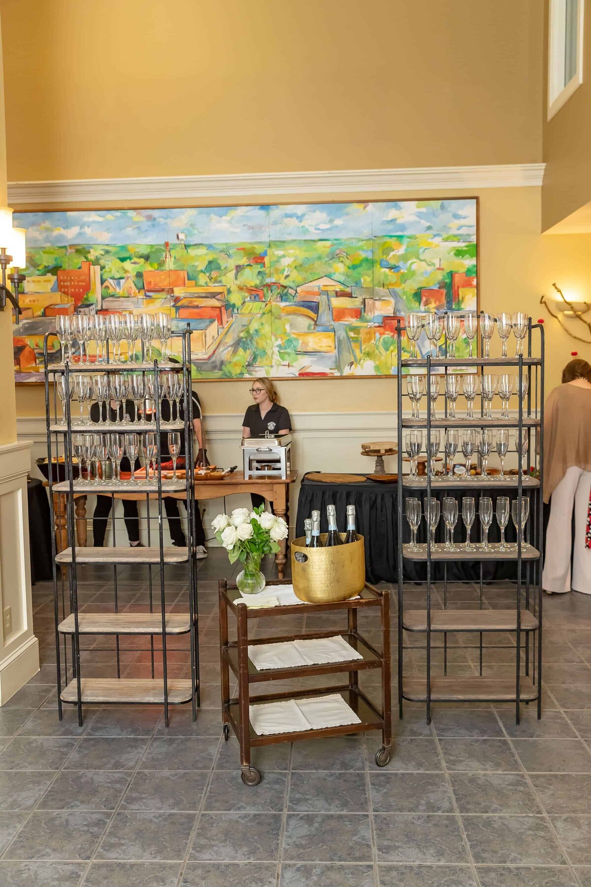 A drink station with racks of empty wine glasses, a cart with a gold ice bucket and bottles, and a person standing behind a table at an indoor event. A colorful painting hangs on the wall in the background.