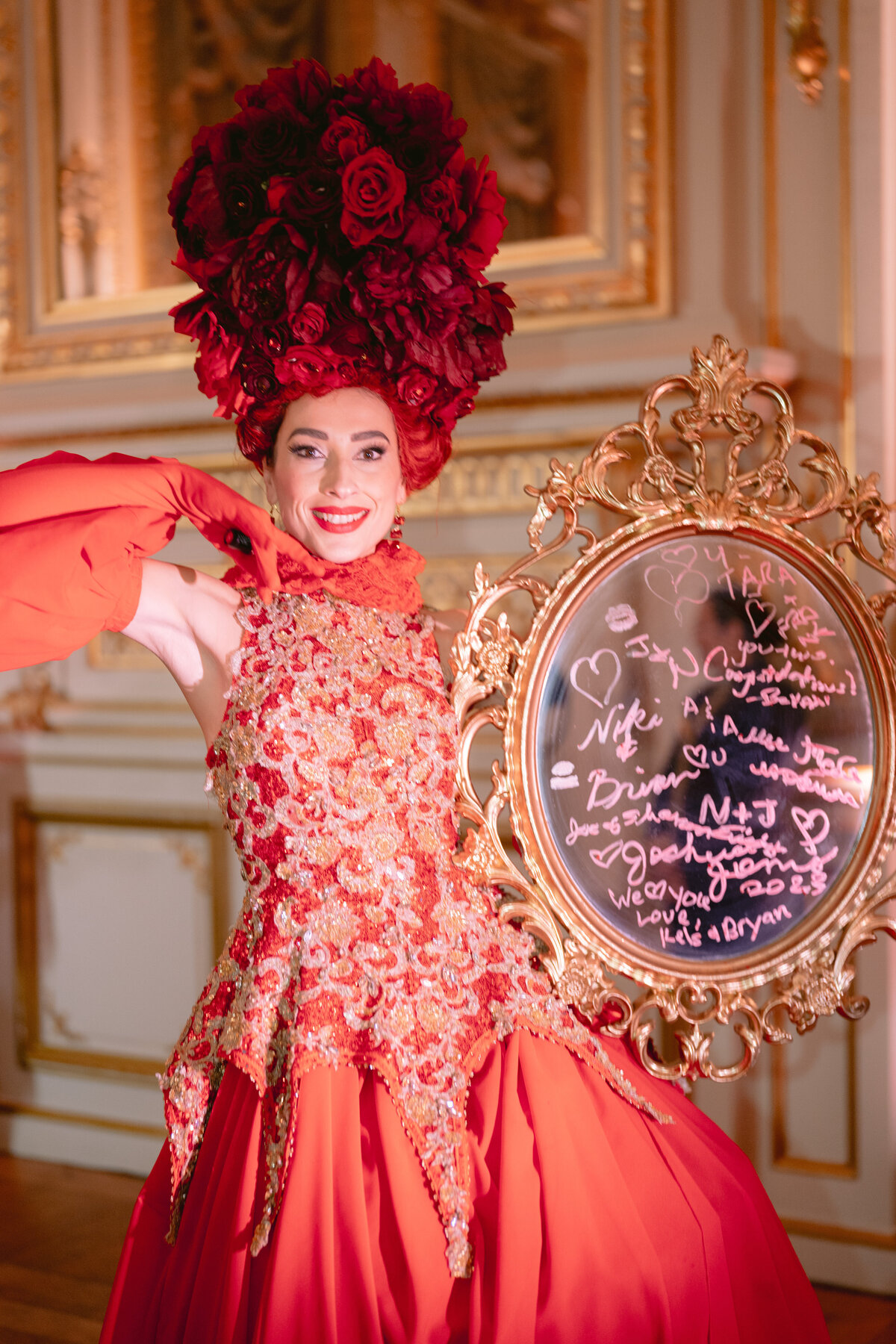 The captivating photo featuring a person dressed in an elaborate red costume adorned with roses and ruffles, wearing a large red flower headdress, and standing in an ornate room with gold details was skillfully captured by photographer Matteo Coltro. Videographers from Zen Film Works contributed to the video footage production of this enchanting scene at the Shangri La Hotel & Ritz Paris Hotel in Paris, France, lending an air of elegance and charm to the overall composition. The individual is depicted with a warm smile, holding a circular frame embellished with various handwritten messages and hearts on the glass, adding a touch of whimsy to the striking imagery.