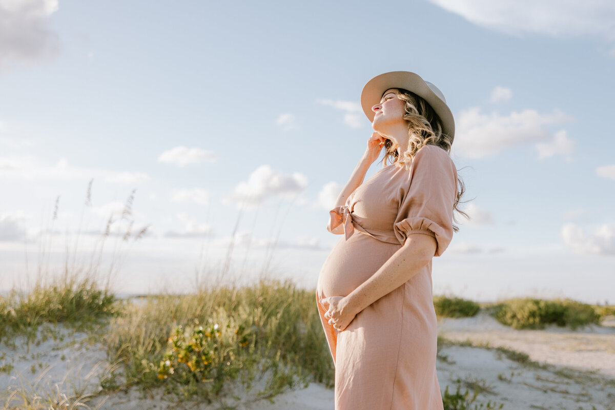 Pregnant woman in pink dress wearing hat on Clearwater beach for Tampa maternity photographer Tina Marie Studio.