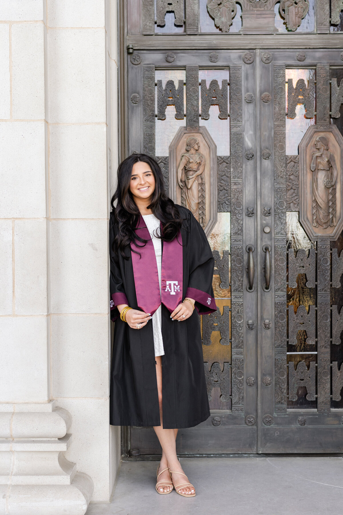 Texas A&M senior girl leaning against front door of Administration Building while smiling and holding maroon stole while wearing gown