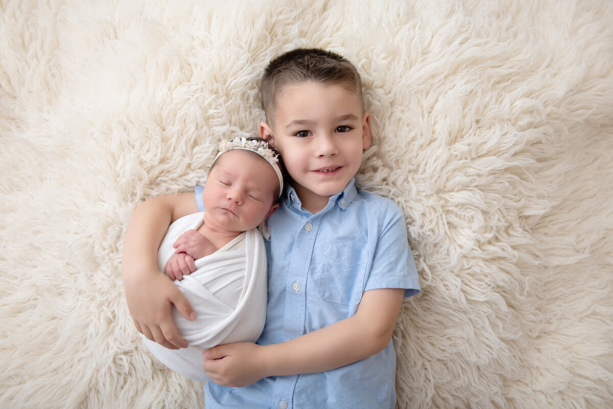 big brother holding little sister laying on a cream rug for their newborn photography session in Burlington , Ontario