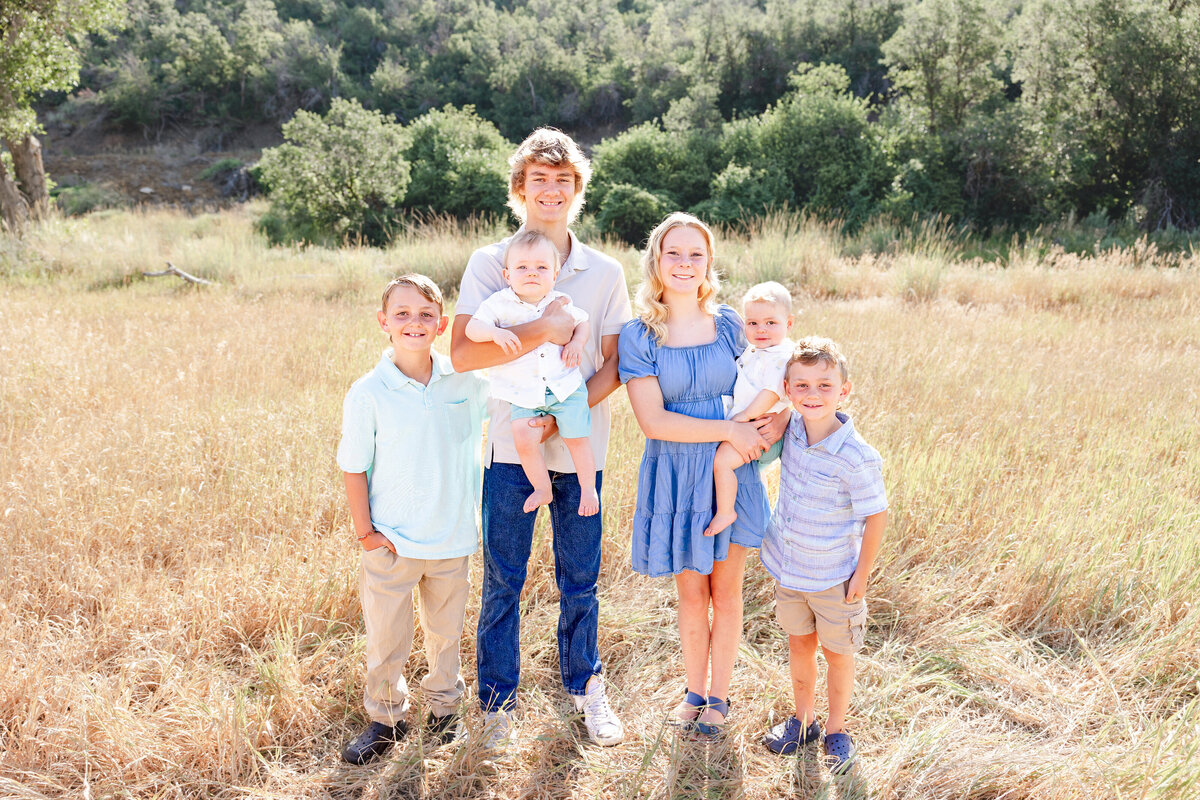 A group of siblings, including older and younger children, standing together in a sunlit field, wearing light-colored outfits. Taken by Cassie Lee Photography