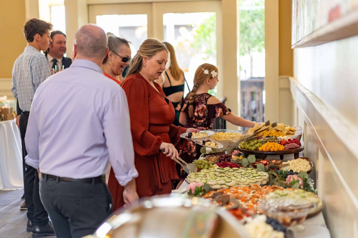 People dressed in semi-formal attire serve themselves food from a buffet table with a variety of dishes, including fruit, crackers, dip, and hot entrees, at an indoor event space.