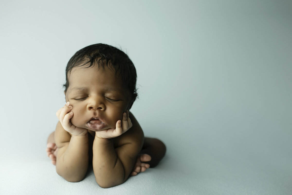 A newborn baby boy posed in the classic froggy position, with his tiny hands gently supporting his chin and his legs tucked under, creating an adorable and serene expression. The baby is wrapped in soft, neutral-toned fabric, resting on a cozy, textured blanket with warm, dreamy lighting enhancing the peaceful atmosphere