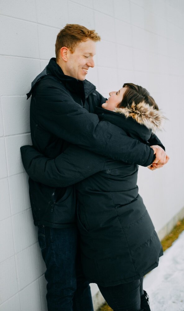 A boyfriend leans against a white wall while embracing his girlfriend