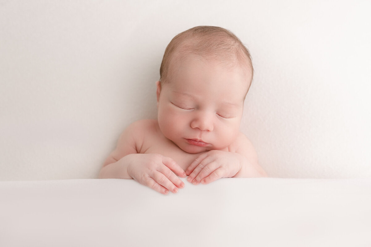Newborn baby sleeping peacefully lying on back and holding onto the blanket that is covering him at the elbows. Baby is lying on a white blanket and covered up by a white blanket at Oregon newborn photography session.