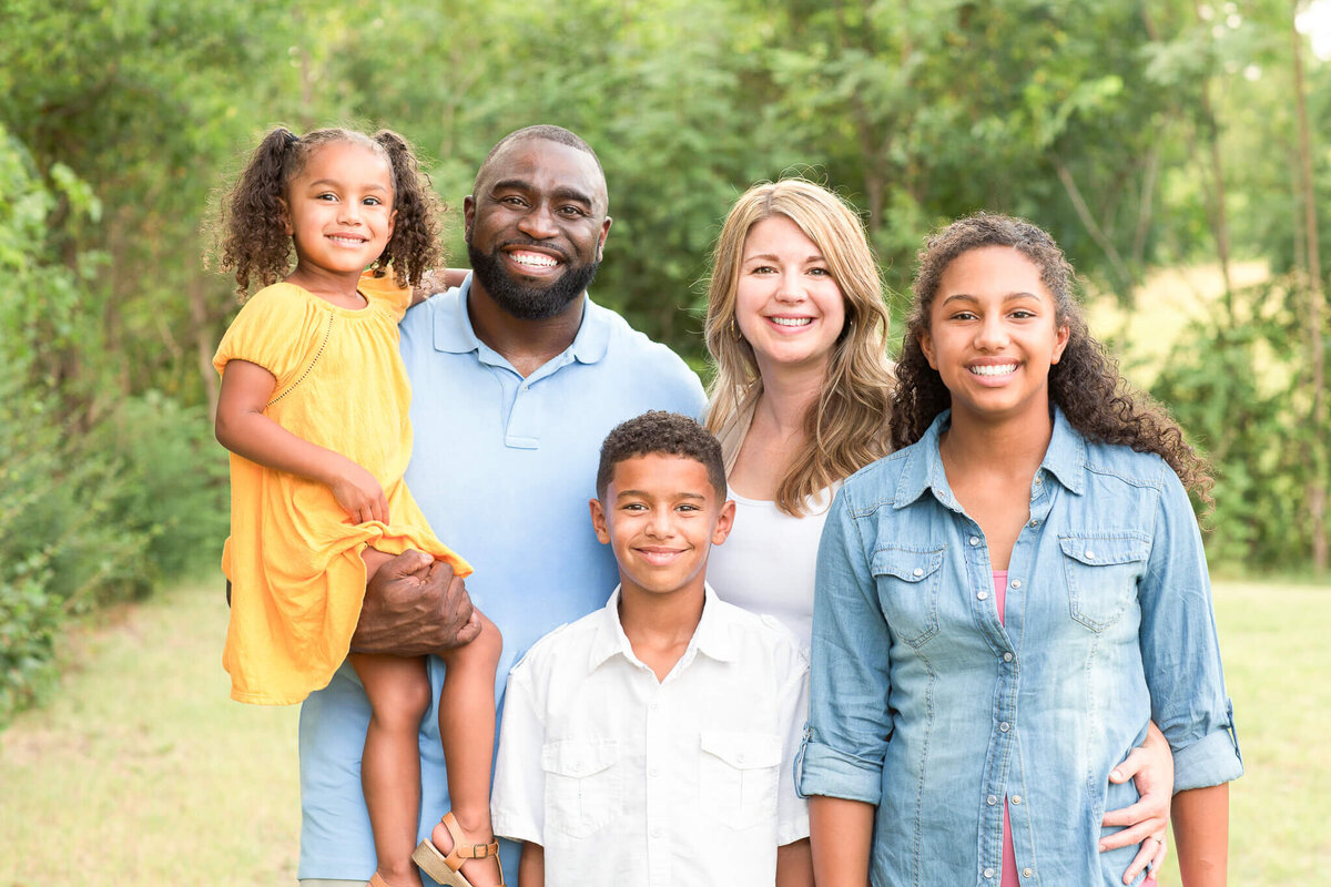 A Fayetteville NC family photographer captures a joyful family of five posing outdoors, all smiling at the camera. The father holds a young girl in a yellow dress, while the mother stands beside two other children—a boy and a teenage girl—set against a backdrop of lush trees and greenery.