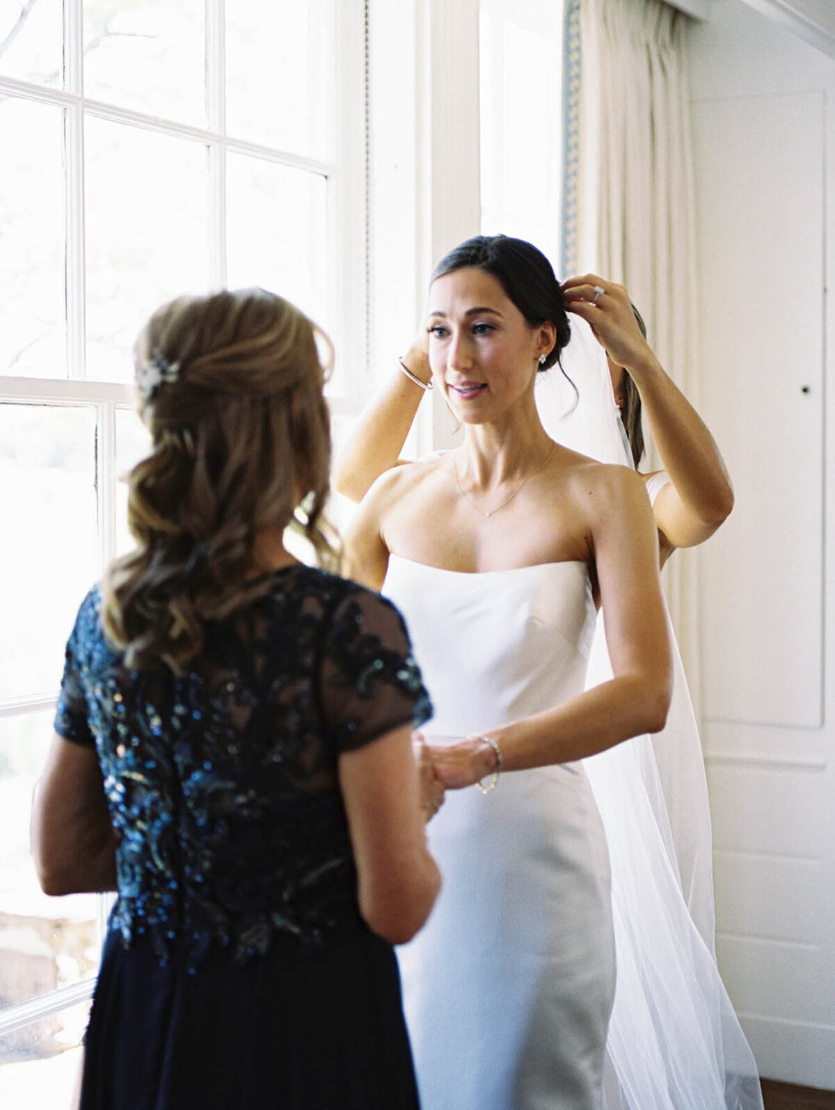 Bride gets her veil put on while she holds her moms hands.