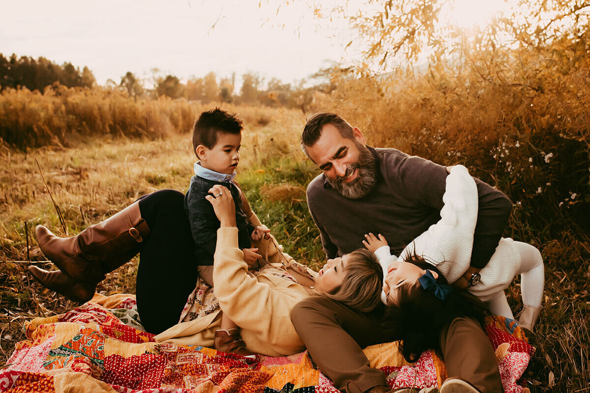 a family playing and laughing together on a quilted blanket at sunset in a field