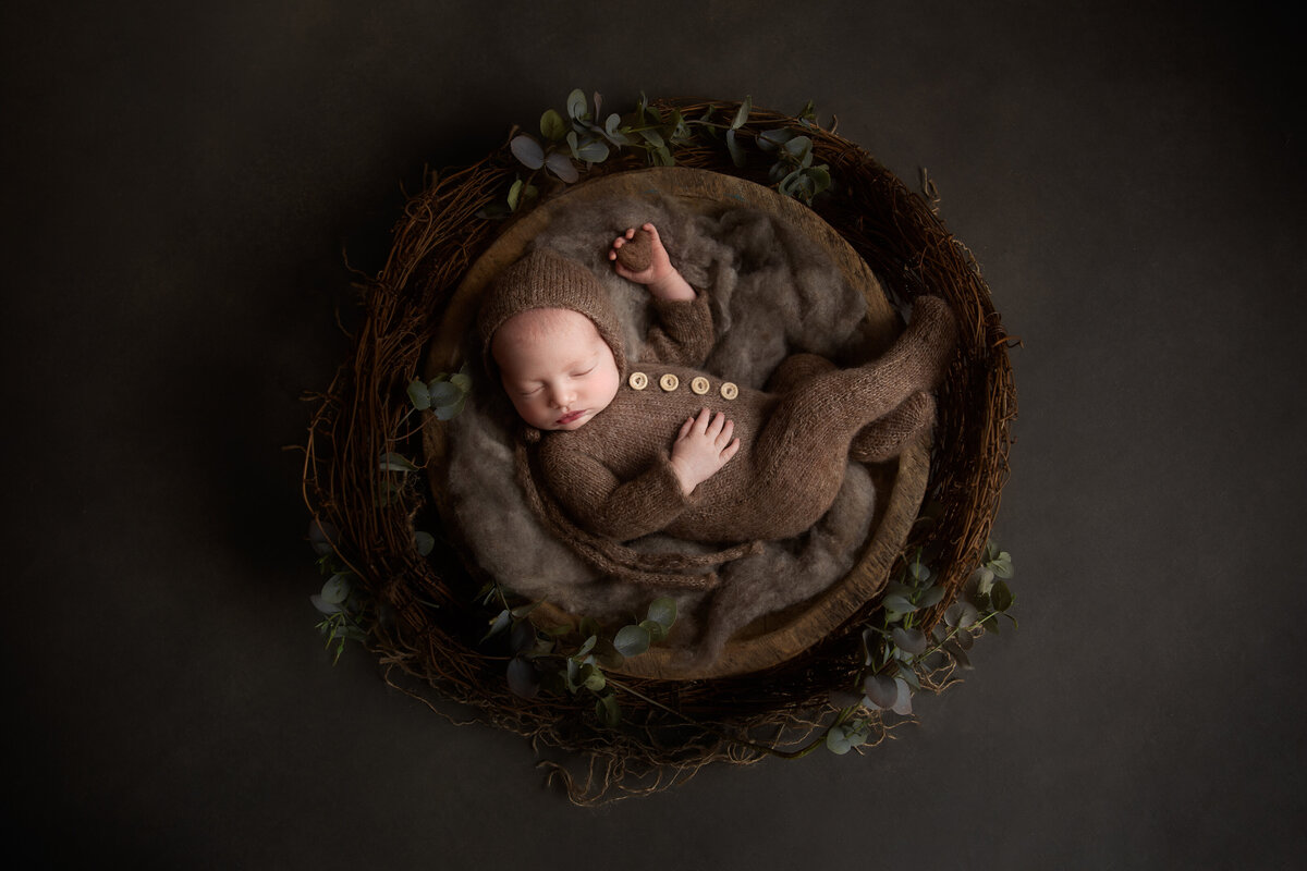 Newborn baby wearing a brown knitted onesie sleeping on her back in a wooden bowl on a grey background holding a little heart in her hand.