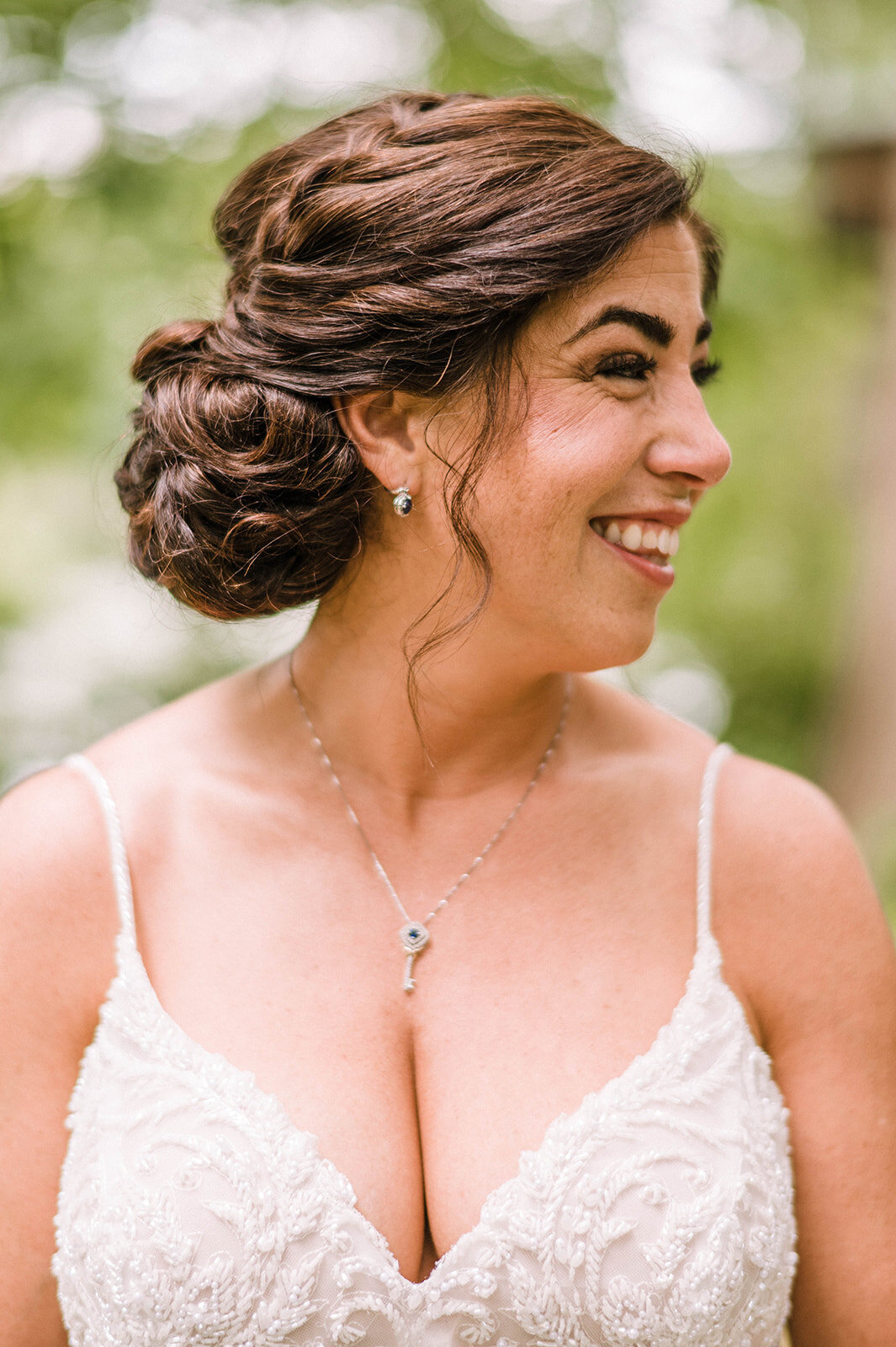 A bride with her hair in an updo wearing a white dress with a key necklace smiles at the Woodlands at Algonkian