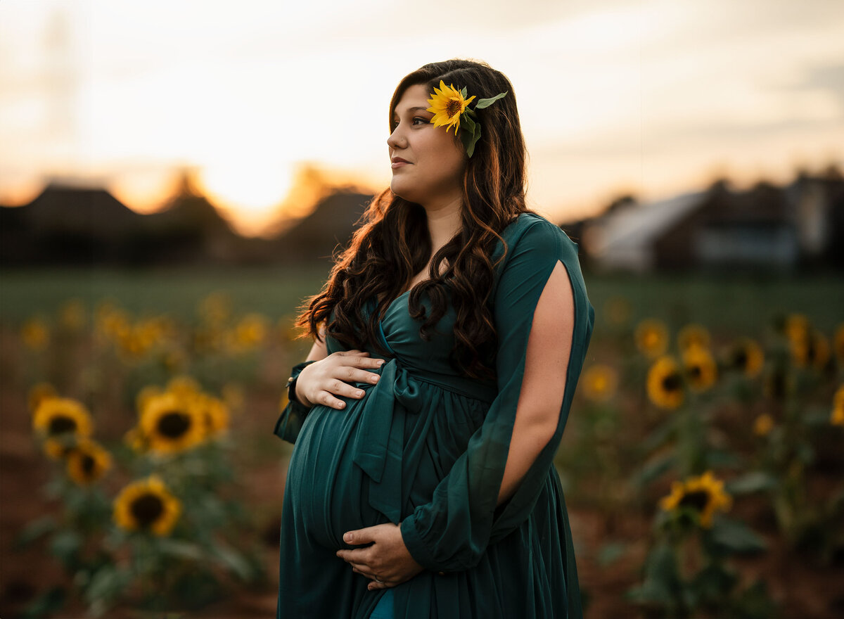 Woman in dark green dress standing amongst a field of sunflowers holding her pregnant belly and looking out at the sunset at sirmon farms