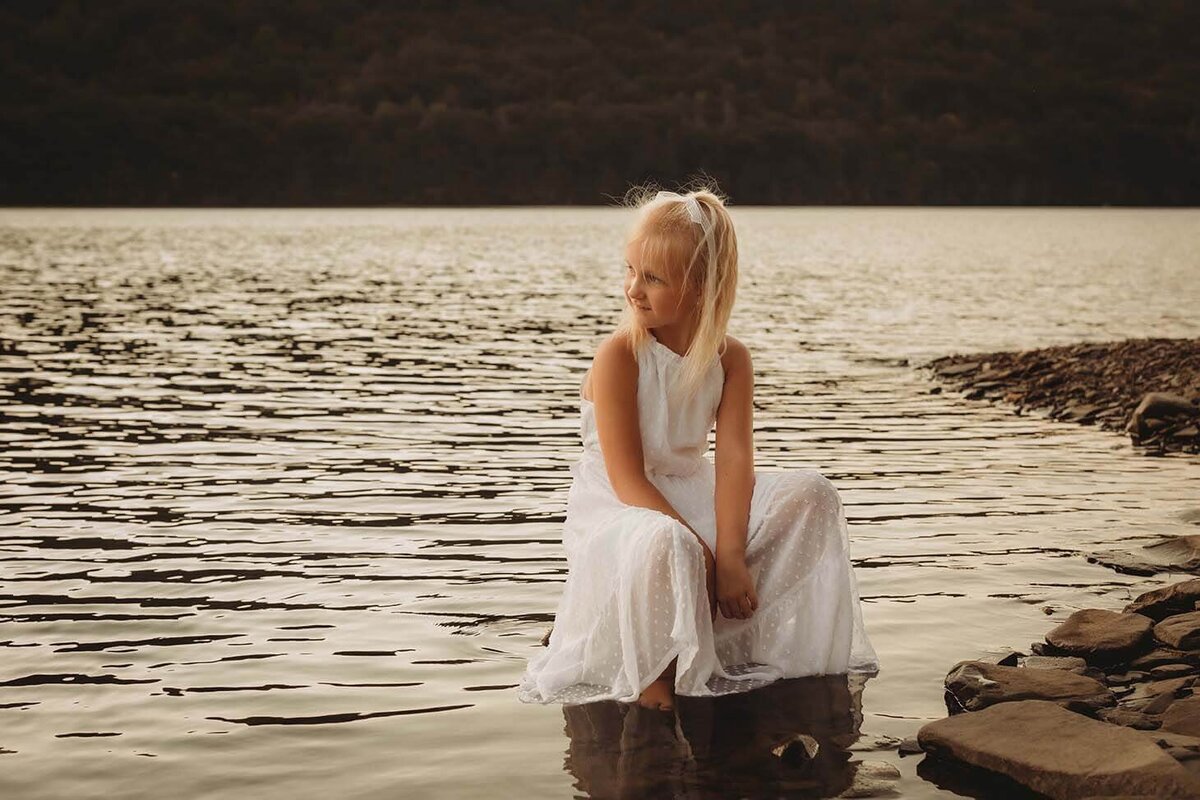 a young girl in a white dress sits on a rock in the center of a lake while looking off in the distance