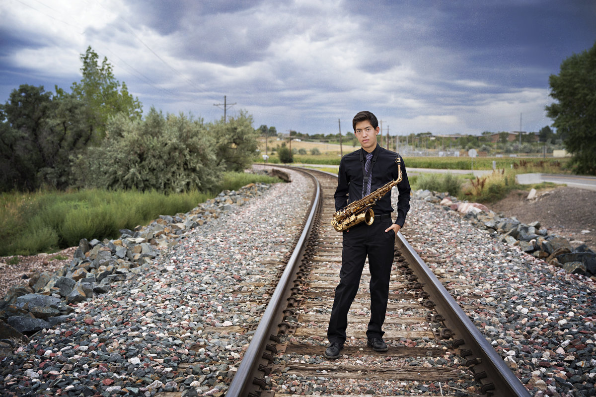Guy poses under a moody sky with his sax and railroad tracks