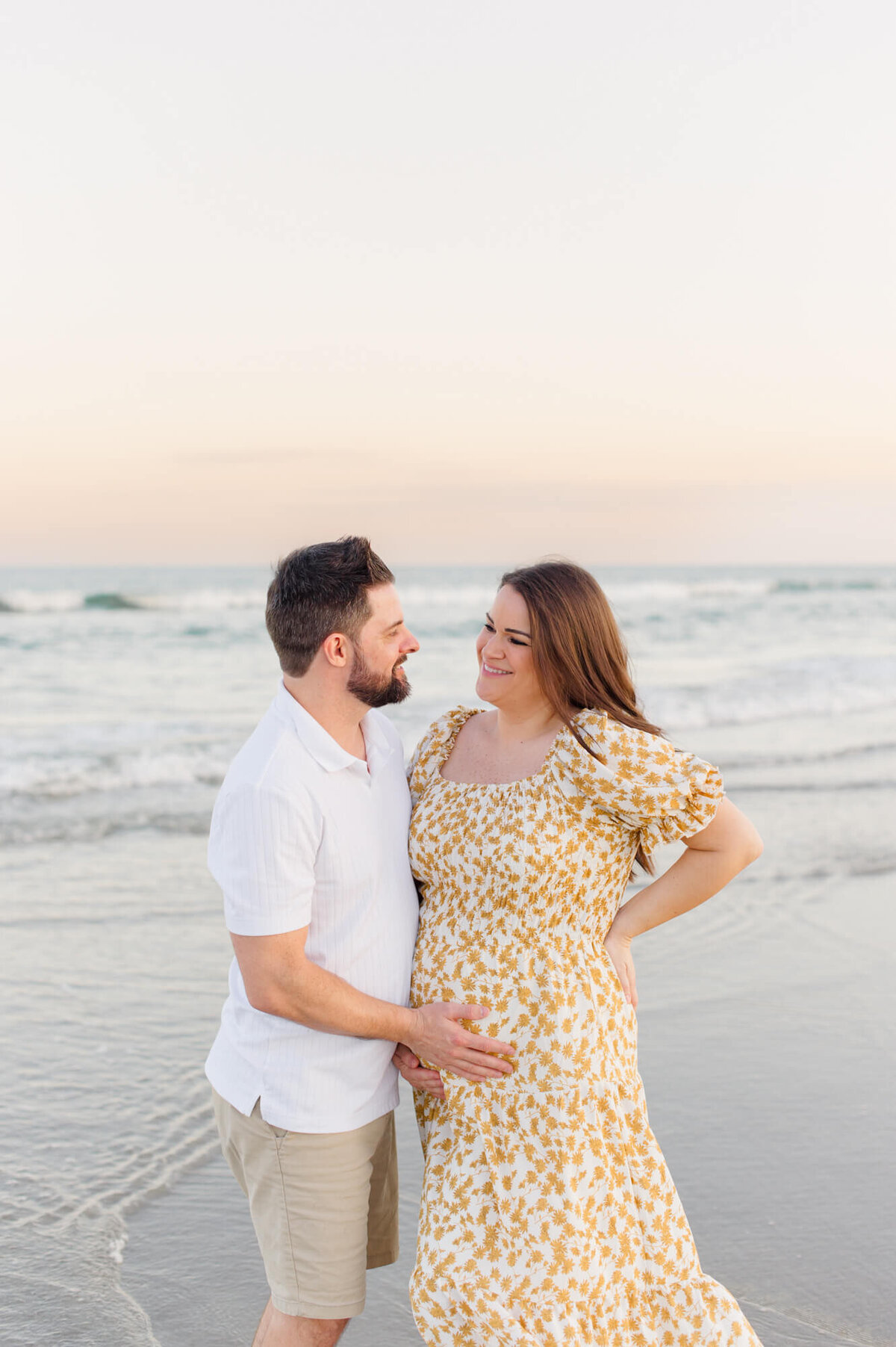 New parents stand on the beach holding mothers belly during their session with an Orlando maternity photographer