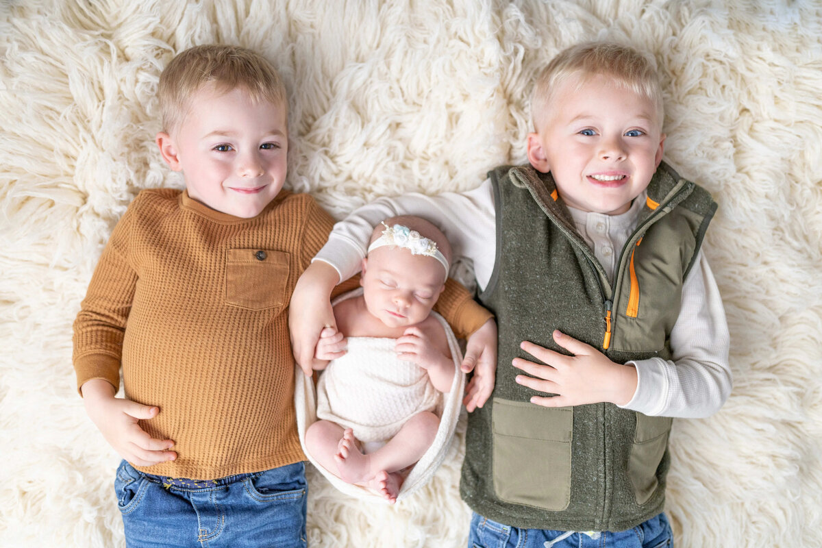 Two brothers cuddling newborn baby girl on cream fur - cozy sibling photo in Maple Grove newborn studio