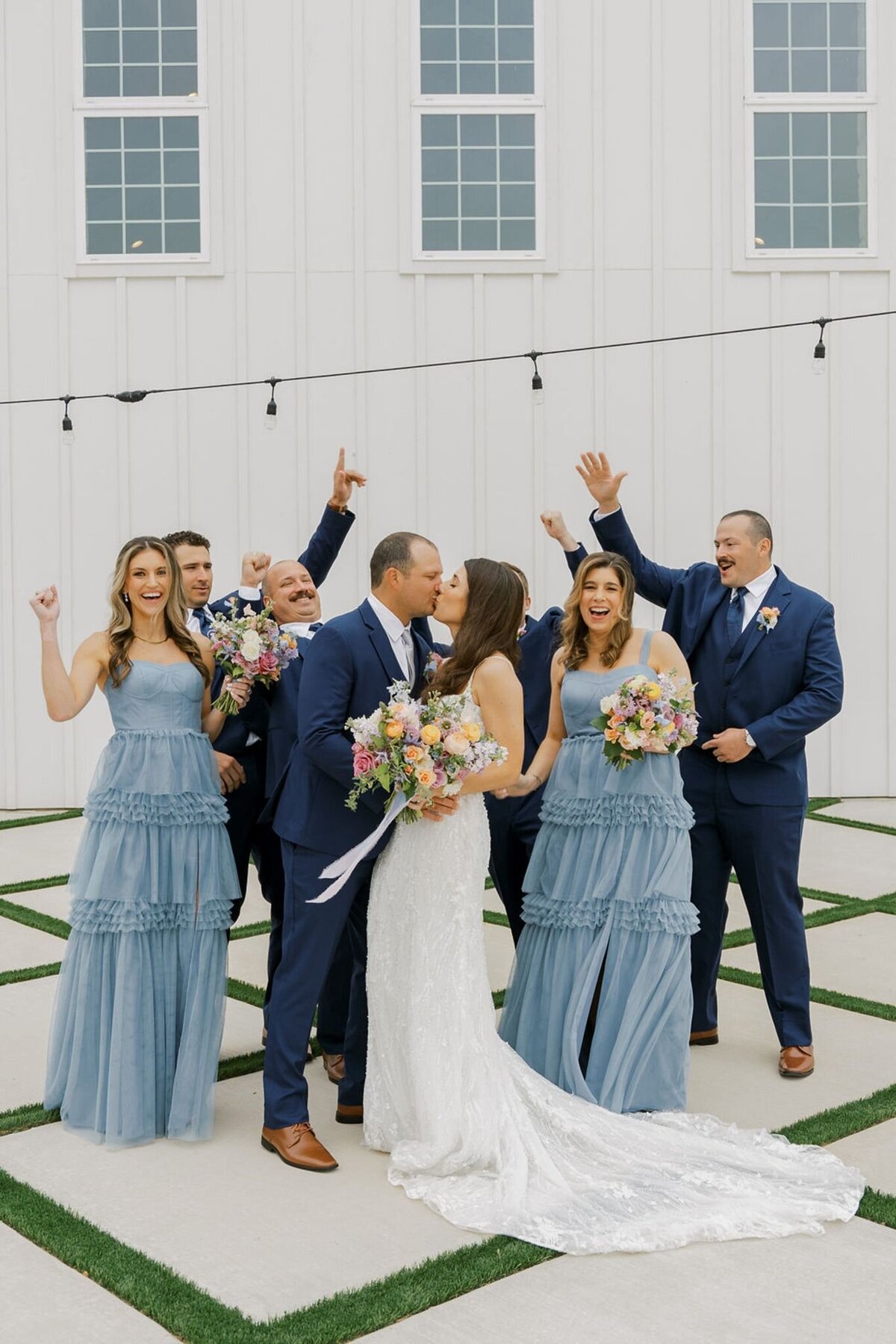 Bride and groom kissing with joyful bridal party, pastel floral bouquets and blue bridesmaid dresses.