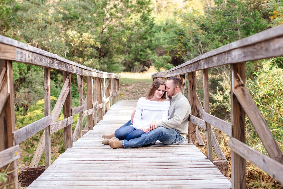 wedding engagement session  on bridge with white sweater couple sitting he whispers she smiles by Firefly Photography of New Braunfels Texas