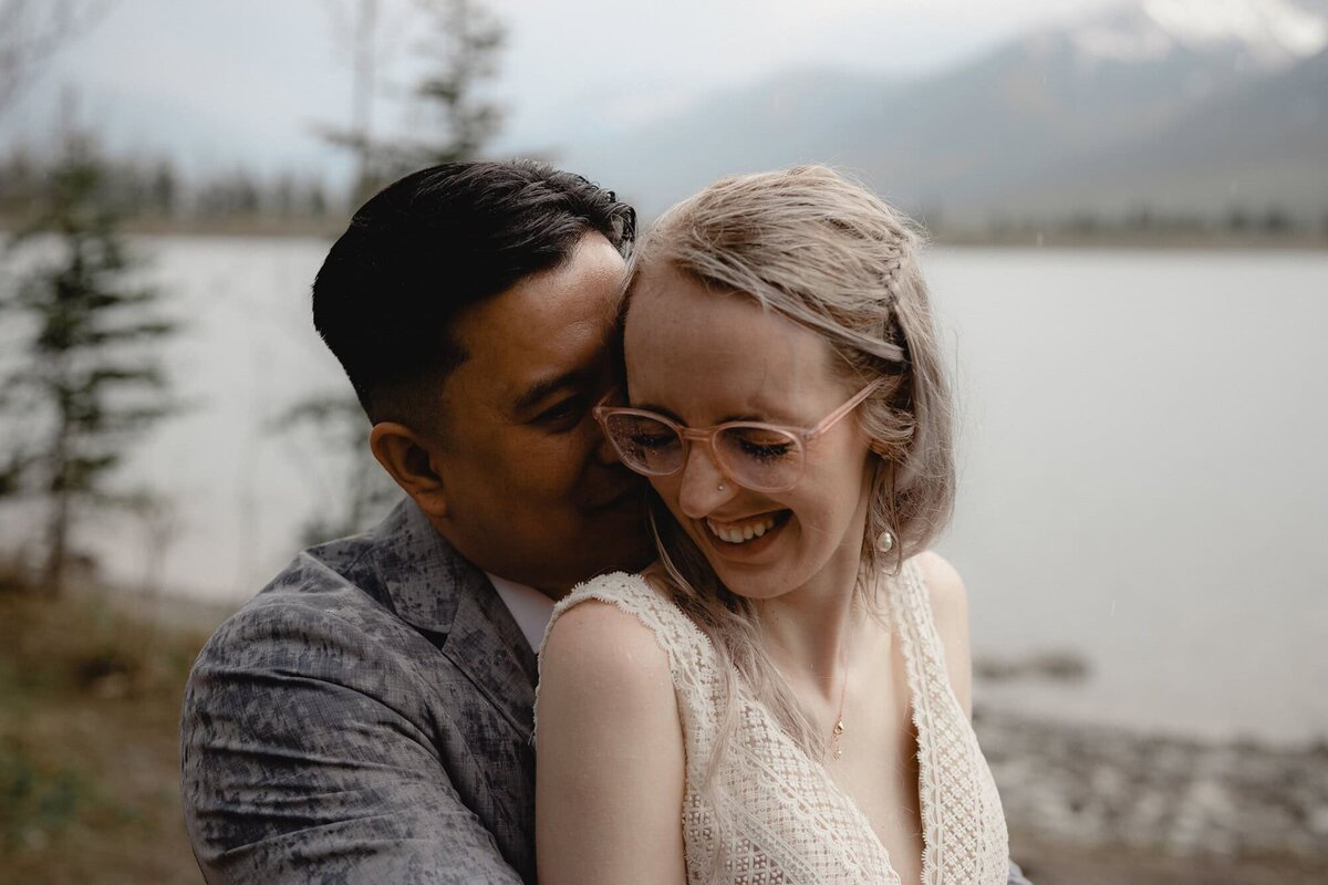 Wedding couple hold each other and smile in front of mountain lake